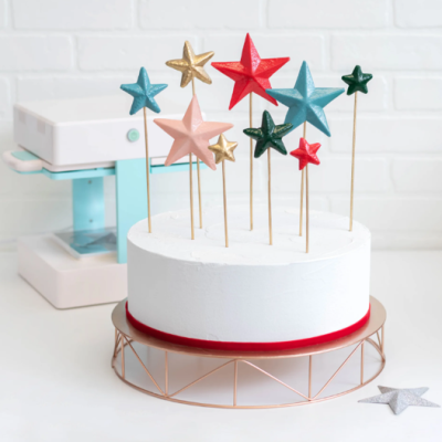 Bolo branco decorado com estrelas coloridas em palitos numa base metálica rosada.