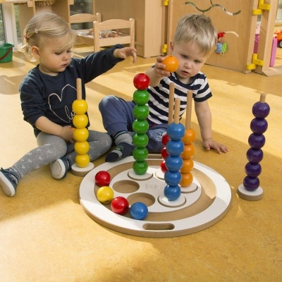 Crianças a brincar com brinquedo educativo de madeira com bolas coloridas empilhadas em pilares numa base redonda no chão.