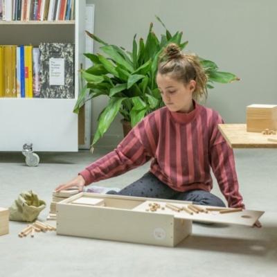 Criança brincando com peças de madeira no chão junto a planta e estante de livros