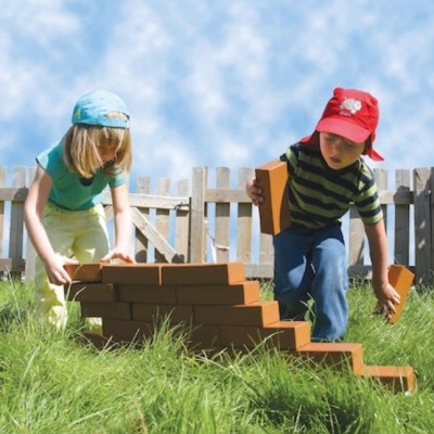 Duas crianças a brincar com blocos laranja na relva perto de uma vedação branca.