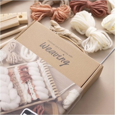 Kit para tecelagem com fios de lã em bege, castanho e branco e caixa de cartão com texto Weaving.