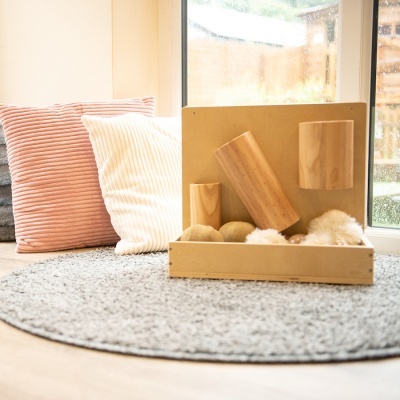 Objetos decorativos em madeira clara sobre tapete cinza com almofadas listradas ao fundo perto de janela