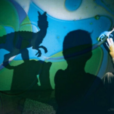 Sombras de crianças e dinossauro projetadas em parede azul e verde