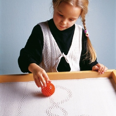 Criança a fazer desenhos com bola de textura em areia numa caixa de madeira
