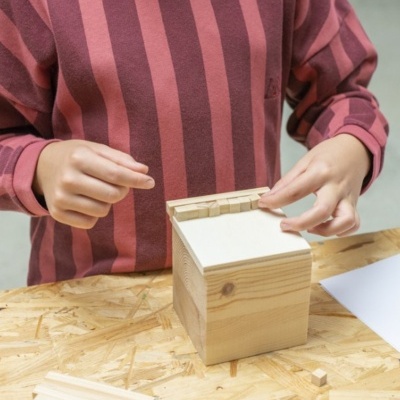 Pessoa a montar caixa miniatura de madeira clara sobre mesa de madeira OSB