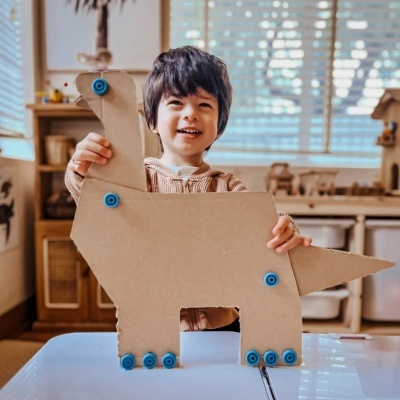 Criança sorridente com figura de dinossauro de cartão castanho com detalhes azuis, em ambiente interior com móveis e janela