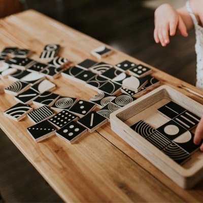 Peças de madeira quadradas pretas e brancas com padrões geométricos sobre mesa de madeira clara, com mão infantil e caixa de madeira