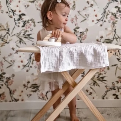 Criança a usar ferro de passar roupa sobre tábua de madeira com papel de parede floral ao fundo