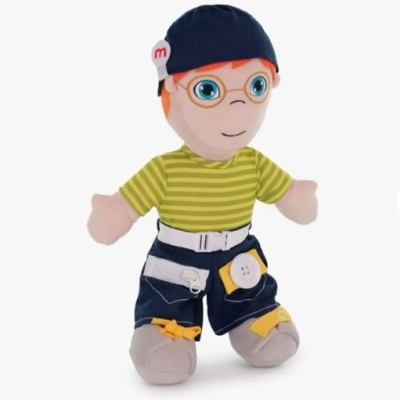 Boneco de peluche vestido com camisa às riscas verdes e amarelas, calções azul escuro e gorro azul.