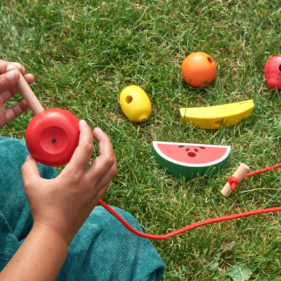 Brinquedos de madeira coloridos com formato de frutas e bolas laranja sobre relva, criança segura peça vermelha redonda com cordão.