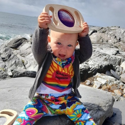 Bebé com roupa colorida e casaco cinza segurando brinquedo de madeira em ambiente rochoso à beira-mar