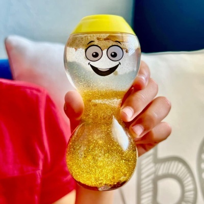 Frasco de plástico transparente em forma de ampulheta com rosto sorridente, tampa amarela e líquido dourado brilhante