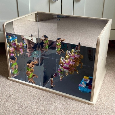 Caixa de madeira com espelhos interiores refletindo figuras de brinquedo coloridas num tapete bege