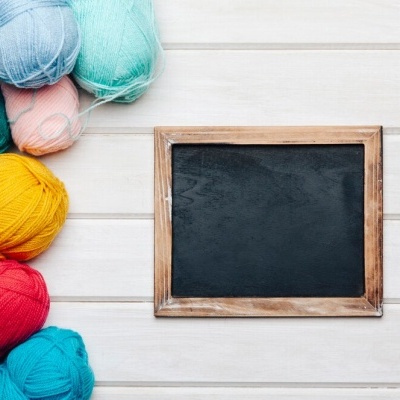 Novelos de lã colorida junto a uma pequena lousa preta com moldura de madeira em fundo branco