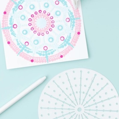 Dois modelos plásticos para desenho circular, um quadrado com padrões rosa e azul e outro redondo azul claro, com penas brancas sobre fundo azul claro.