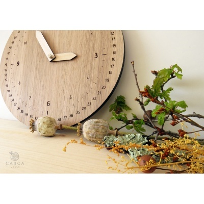 Relógio de parede de madeira clara com números escuros e ponteiros de madeira, junto a flores secas e ramos na base