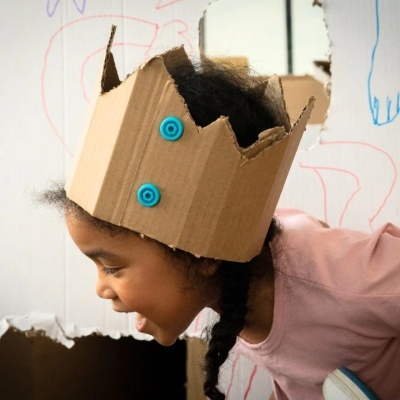 Criança com coroa de cartão bege e rodas azuis, camisa rosa, em ambiente com paredes brancas e desenhos coloridos.