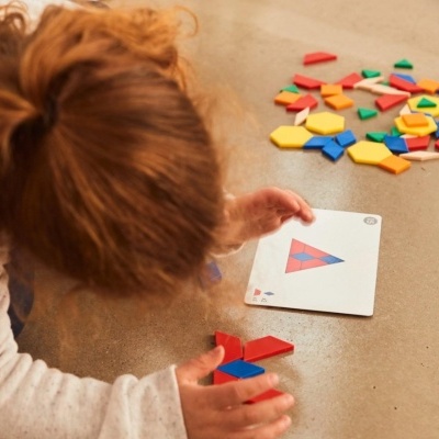 Criança a montar padrão triangular com peças geométricas coloridas e cartão de instrução no chão.