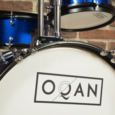 Bateria acústica azul com pele branca e logótipo OQAN montada em sala com parede de tijolos
