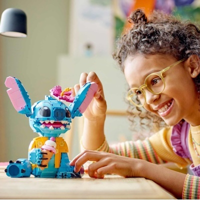 Menina com óculos amarelos brinca com figura de brinquedo azul de Stitch em mesa de madeira.