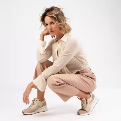 Mulher com camisa bege clara, calças rosa claro e sapatilhas bege sobre fundo branco