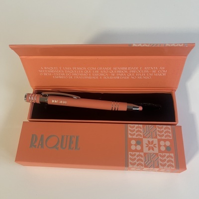 Caneta coral numa caixa coral com o nome RAQUEL e padrão floral, texto em português na tampa da caixa.