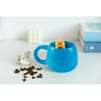 Caneca azul com figura de pato amarelo, grãos de café e biscoitos ao lado, livros azuis desfocados ao fundo