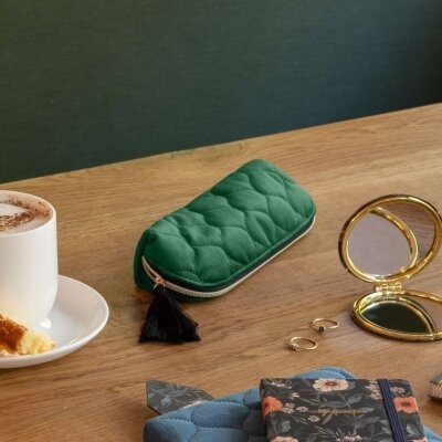 Estojo verde almofadado com fecho dourado e pendente preto sobre mesa de madeira com espelho compacto dourado, folhas decorativas, anéis dourados e chávena de bebida com bolo