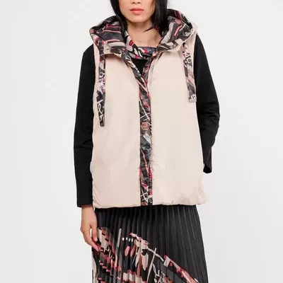 Colete acolchoado bege com padrão abstrato em capuz e fecho, usado com blusa preta e saia plissada colorida.