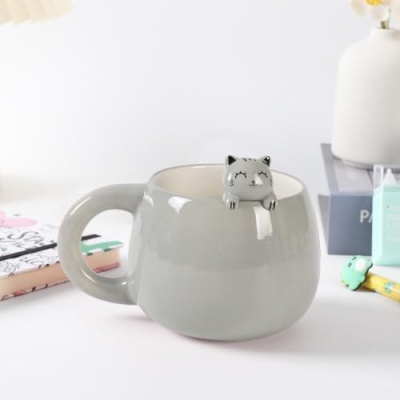 Caneca cinza com figura de gato pendurada na borda em mesa branca