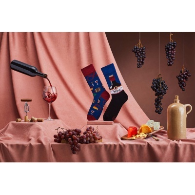 Meias com padrão de vinho junto de copo a ser servido, uvas, queijo e jarro em fundo cor de rosa e castanho