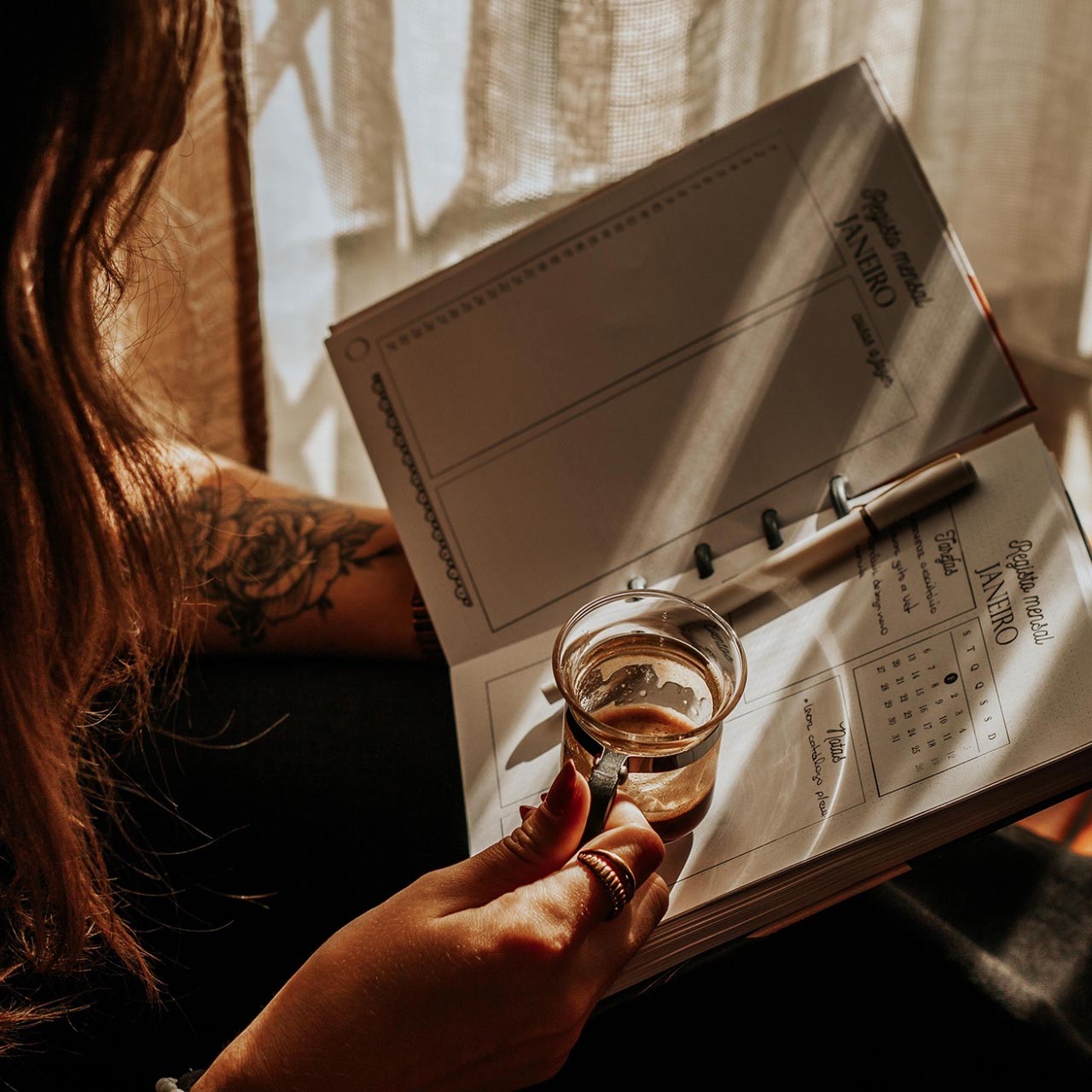 Agenda aberta com caneta e copo de café nas mãos de uma pessoa junto a uma cortina iluminada pela luz do sol.