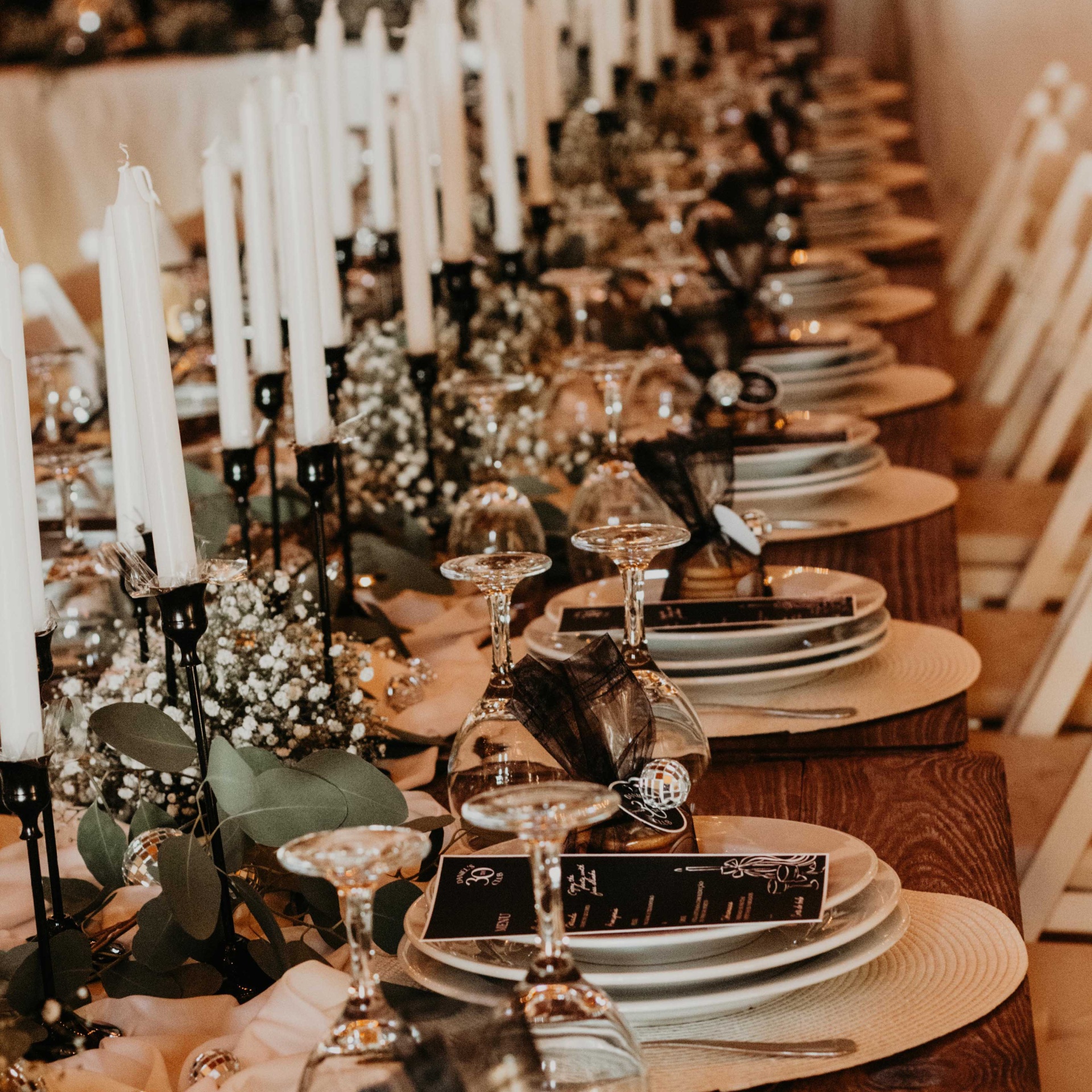 Mesa de jantar decorada com velas brancas, pratos e copos, menus e flores