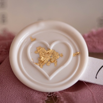 Selo branco redondo com relevo em forma de coração e fragmentos dourados no centro sobre tecido rosa.
