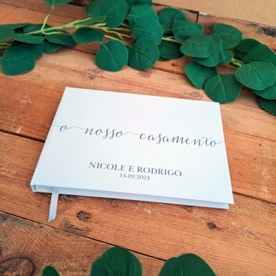 Livro de assinatura branco para casamento com texto personalizado, sobre mesa de madeira com folhas verdes