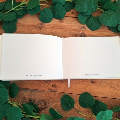 Livro aberto com páginas em branco, texto 'NICOLE E RODRIGO', sobre mesa de madeira com folhas verdes ao redor