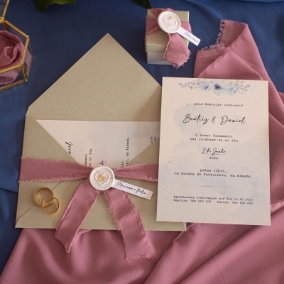 Convite de casamento com envelope bege e fita rosa desfiada sobre tecido azul e rosa.