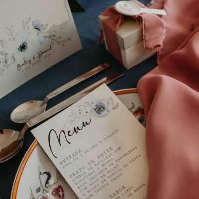 Mesa posta com prato decorado, talheres, menu em português, caixa de presente bege com fita e cartão de convite