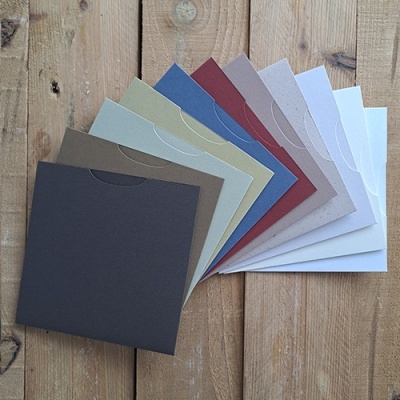 Conjunto de 10 envelopes quadrados em tons neutros e pastel, dispostos em leque sobre madeira.