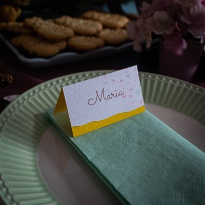Cartão de mesa com nome Maria sobre guardanapo verde claro em prato branco e verde, com doces e flores ao fundo