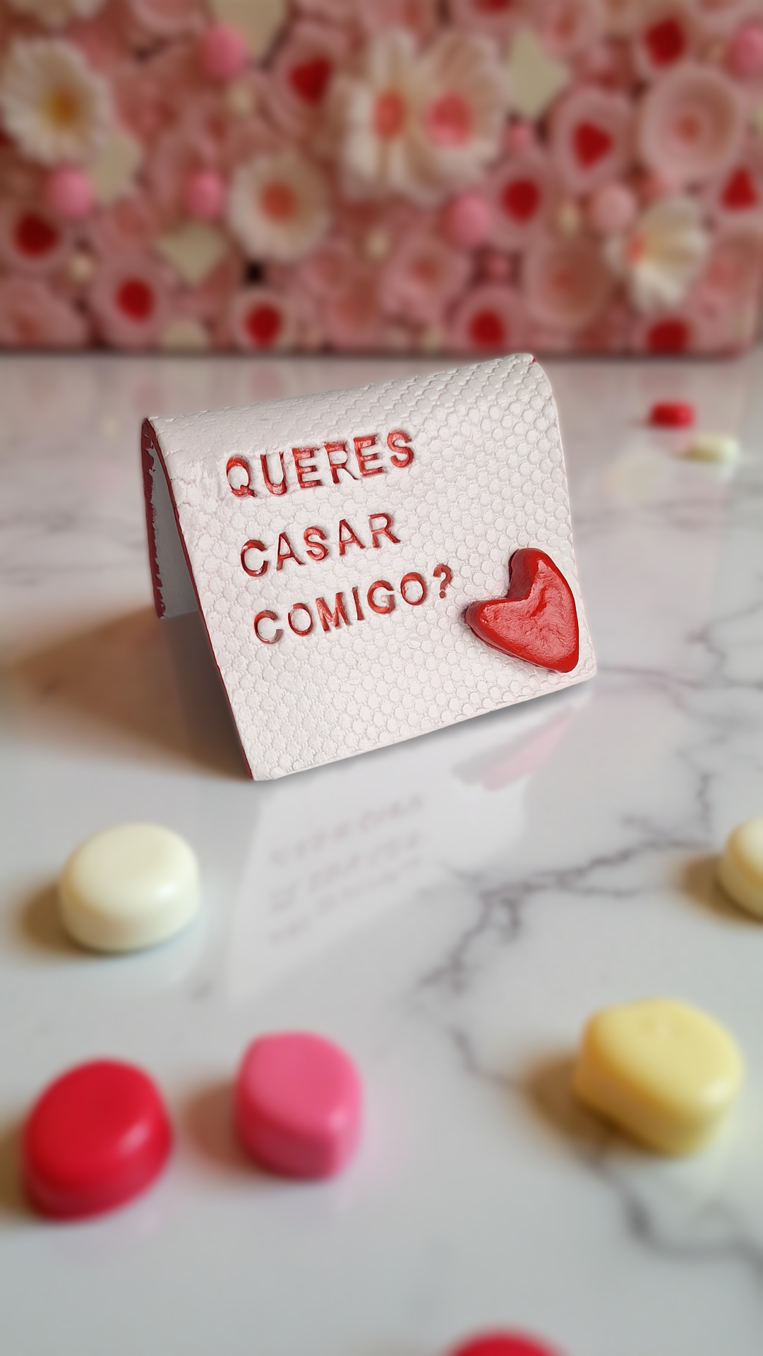 O Mensageiro | Peça Personalizada em Cerâmica Fria Artesanal Quadro branco com mensagem de proposta de casamento em relevo vermelho e coração vermelho sobre mesa de mármore com doces coloridos