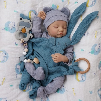 Bebé vestido com fato cinzento de algodão a dormir segurando brinquedo coelho azul e mordedor de silicone e madeira sobre manta branca com estampados