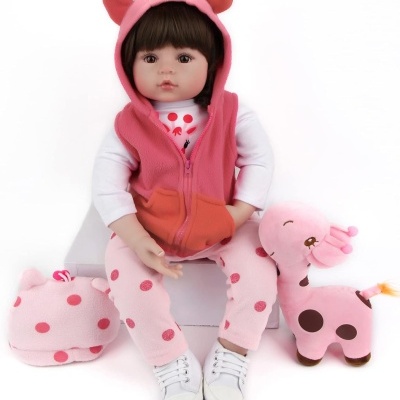 Boneca de roupa rosa com capuz com orelhas, calças cor de rosa com bolinhas, sentada entre bolsa de tecido rosa com bolinhas e peluche de girafa rosa.