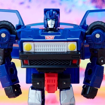 Figura de ação robô azul com detalhes coloridos e símbolo Autobot em fundo desfocado