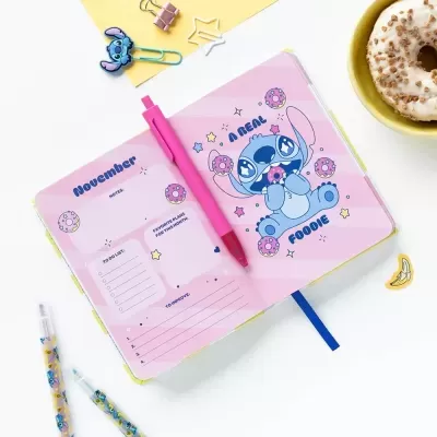 Agenda rosa aberta com personagens Stitch, caneta cor-de-rosa, lápis decorados e donut num prato amarelo