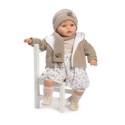 Boneca bebé vestida com casaco bege, gorro e cachecol com pompons, calções florais, sentada numa cadeira branca