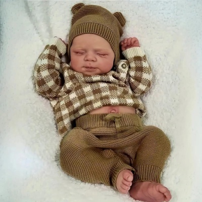 Boneca bebé com roupa de malha xadrez castanha e branca, e gorro castanho a dormir sobre tecido branco.