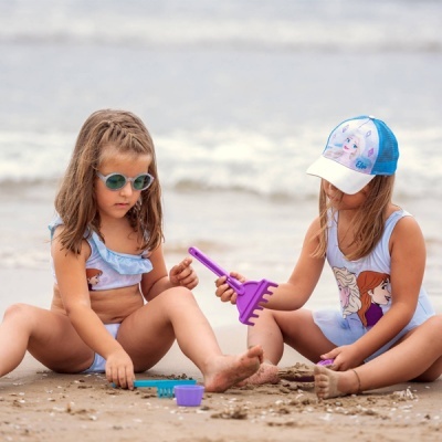 Duas crianças a brincar com brinquedos de areia na praia, fatos de banho com personagens animados, boné e óculos de sol.