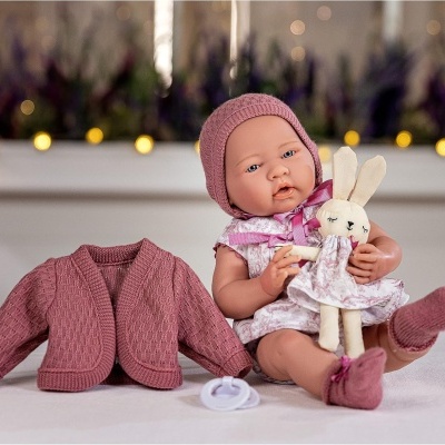 Boneca bebé vestida com vestido branco e rosa e gorro cor de rosa, segurando coelho de peluche, casaco cor de rosa ao lado