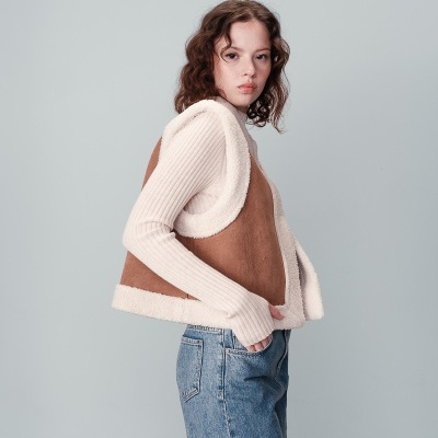 Mulher com colete castanho e branco e camisola de malha branca, calças de ganga azuis, fundo cinzento.